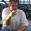 Man pointing, sitting outdoors in casual attire.