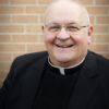 Smiling man in clerical attire, brick background.
